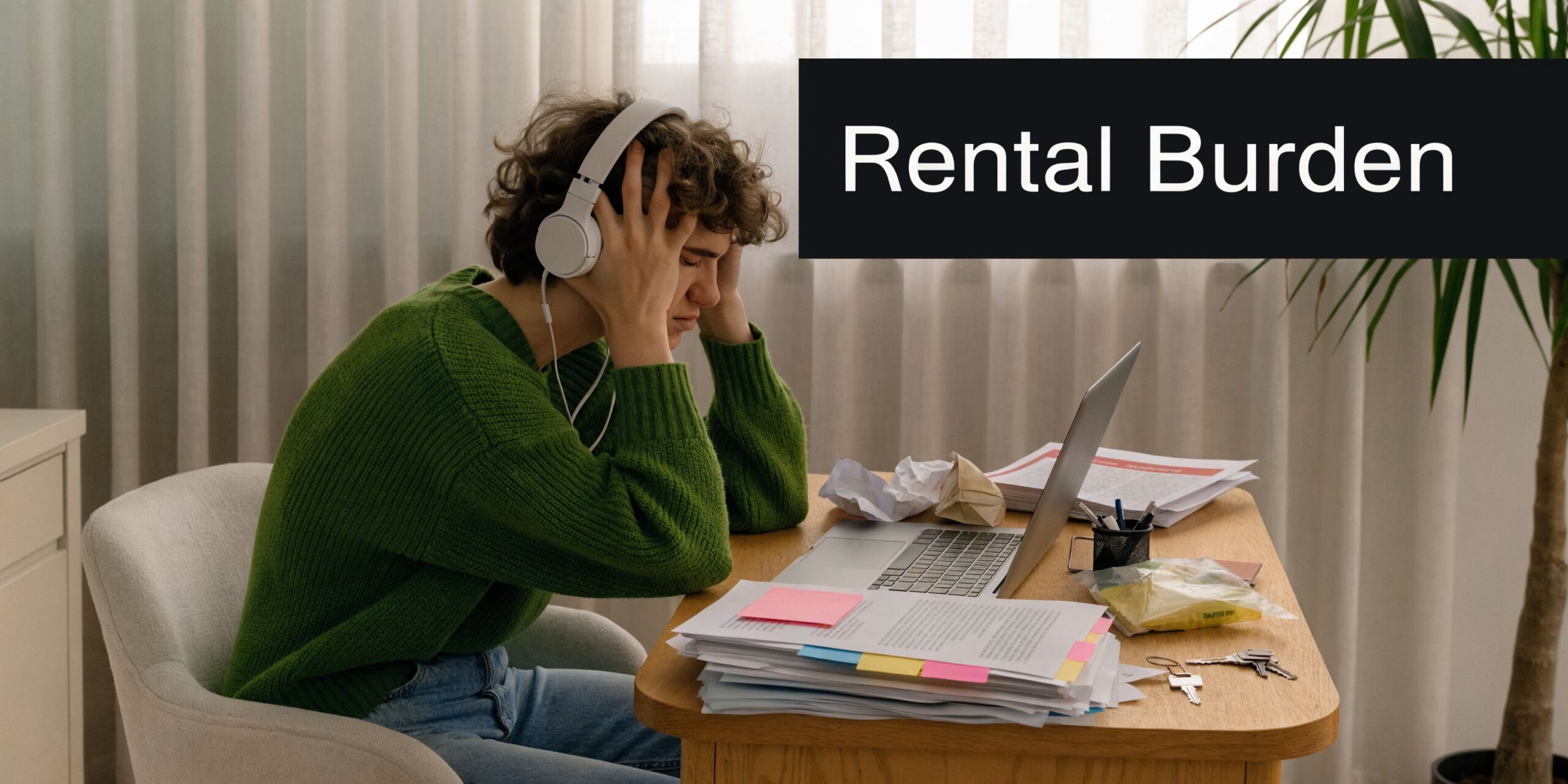 A stressed student wearing headphones sits at a desk with a laptop and piles of paperwork.