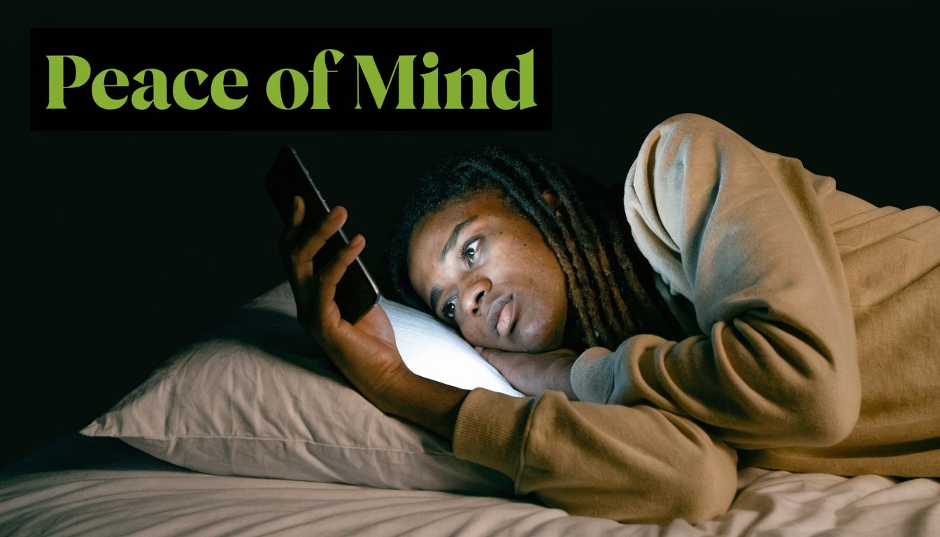 A young person with dreadlocks looks at their glowing smartphone while lying awake in bed at night.
